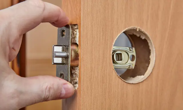 Dłoń wsuwa metalowy mechanizm zamka do wyciętego otworu w drewnianych drzwiach. Zastanawiasz się, ile kosztuje wymiana wkładki w drzwiach?