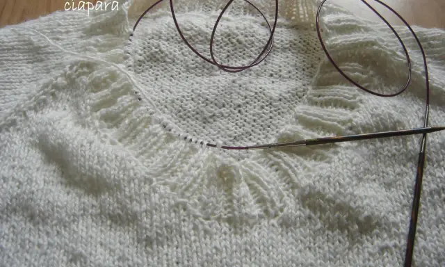 Biały sweter w trakcie dziergania, widoczny fragment kołnierza i druty. Jak zrobić kołnierz do swetra na drutach?