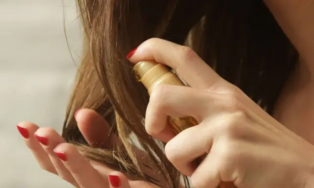 Eine Hand mit roten Nägeln sprüht ein goldenes Serum auf trockenes Haar.