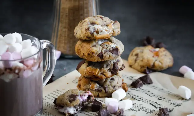 Un cookie chamallow fondant, empilé avec d'autres, à côté d'une tasse de chocolat chaud garnie de guimauves.