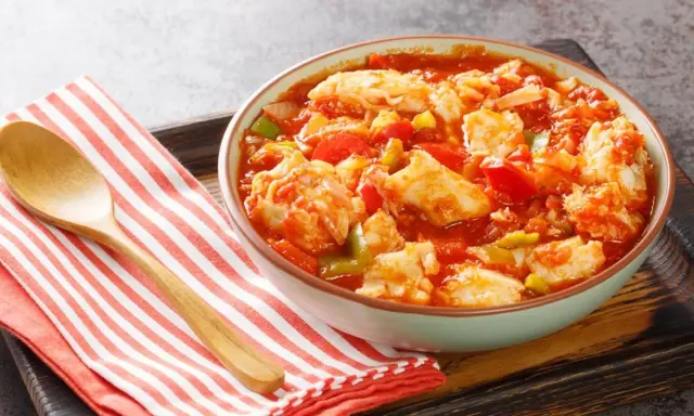 Un tazón de bacalao ajoarriero con pimientos y tomate, servido con una cuchara de madera sobre una servilleta a rayas.