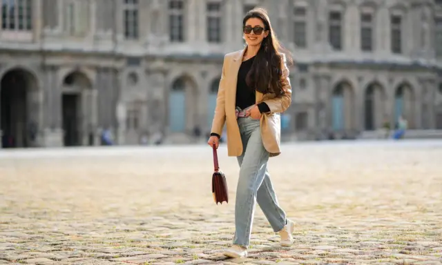 Eine Frau mit langen Haaren und Sonnenbrille spaziert lässig in weiten Jeans und einem beigen Blazer vor einer historischen Fassade.