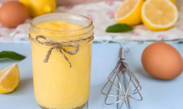 Un tarro de lemon curd casero, atado con cuerda, junto a limones, huevos y un batidor.