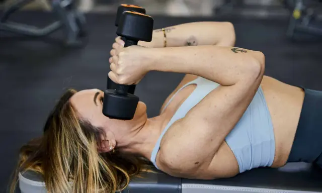 Donna sdraiata esegue skullcrushers con manubri, un esercizio efficace per i gruppi muscolari da allenare insieme, come tricipiti e spalle.