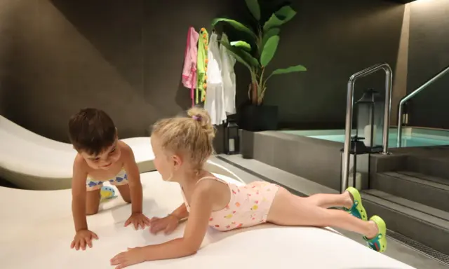 Niños jugando en un spa para niños, con una piscina al fondo y toallas colgadas.