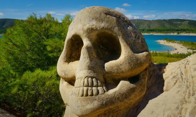 Escultura de calavera en la **ruta de las caras**, con un lago azul y colinas verdes al fondo.