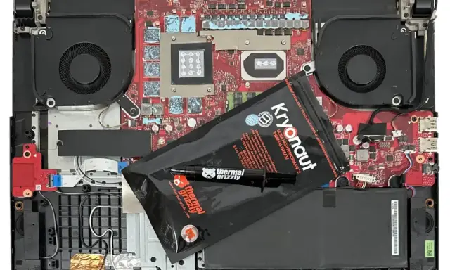 Wnętrze laptopa z wentylatorami i płytą główną, gotowe do wymiany pasty termoprzewodzącej. Opakowanie Kryonaut Thermal Grizzly leży na komponentach.