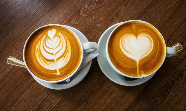 Zwei Cappuccinos mit Latte Art auf einem Holztisch. Sie bestehen aus Espresso, heißer Milch und Milchschaum.