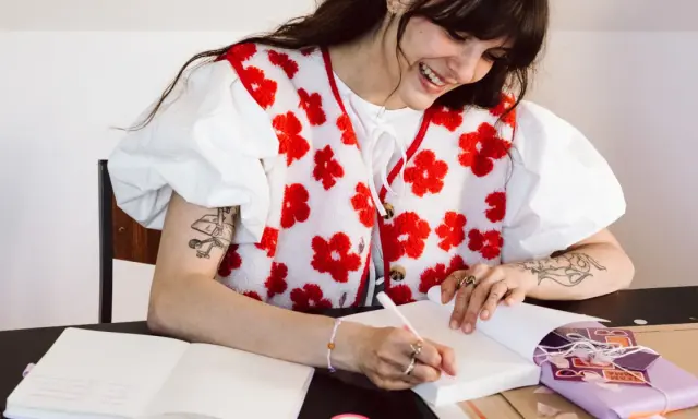 Une jeune femme souriante fait une dédicace livre, stylo à la main, devant un cahier ouvert et un paquet cadeau violet.