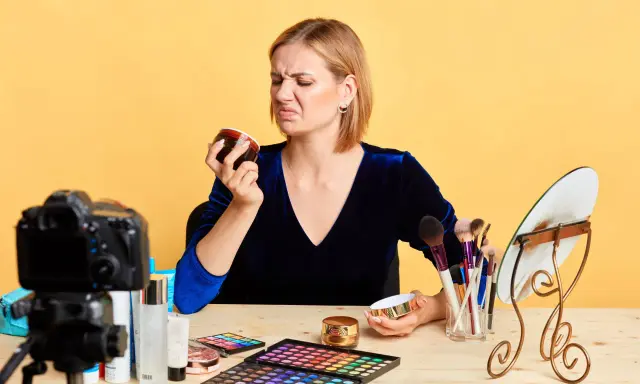 Donna con espressione disgustata annusa una crema viso chiusa, forse per verificarne la scadenza. Sul tavolo, trucchi e una telecamera.