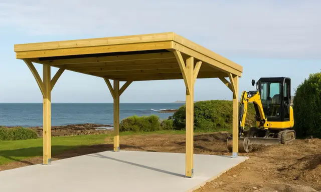 Un carport en bois est en cours de construction près de la mer. Une mini-pelle jaune est garée à côté, prête pour la suite des travaux. Une déclaration de carport est nécessaire pour ce projet.