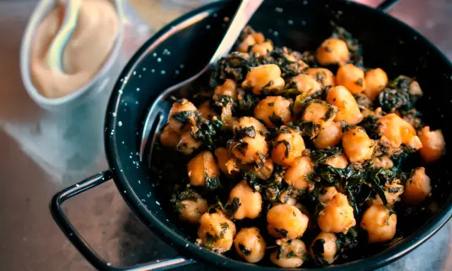 Un plato de garbanzos con espinacas, servido en una sartén negra con un tenedor. Al fondo, una pequeña taza con una salsa cremosa.