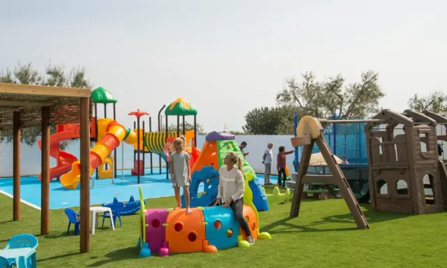 Un parco giochi colorato in una masseria per bambini in Puglia, con scivoli, tunnel e altalene. Famiglie si divertono sotto il sole.