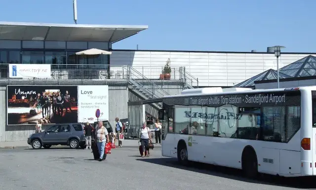 Autobus wahadłowy na lotnisko Oslo Torp. Ludzie z bagażami wychodzą z terminala.