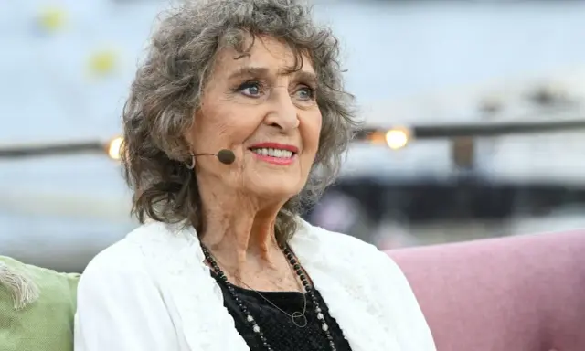 Siv Malmkvist, med sitt karaktäristiska lockiga gråa hår och ett varmt leende, sitter och tittar åt sidan. Hon bär en vit kofta över en svart klänning och smycken.