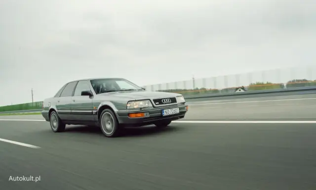 Szary Audi V8 pędzi po autostradzie, chmury na niebie i zielone drzewa w tle.