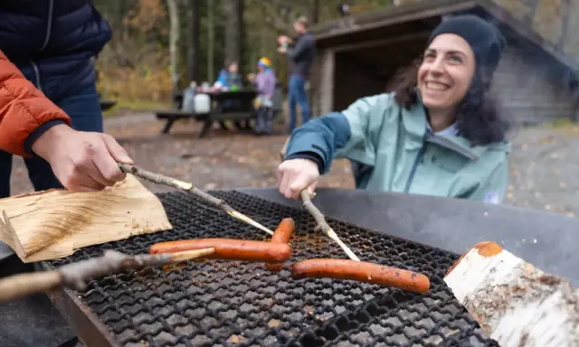 Vänner grillar korv vid en eldstad på Otto Pers gård. En kvinna ler glatt medan hon grillar.