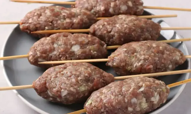 Siedem surowych kofta kebab na patyczkach, ułożonych na szarym talerzu, gotowych do grillowania.