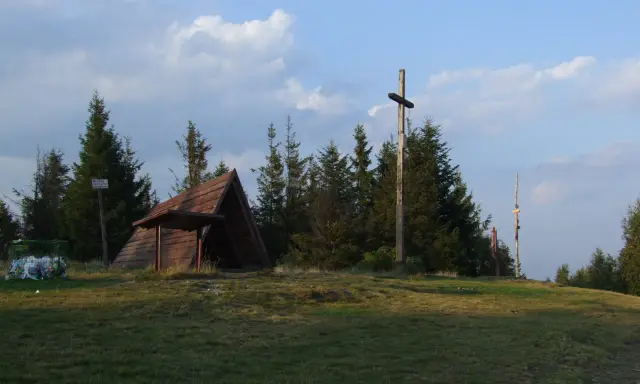 Drewniana wiata, krzyż i kosz na śmieci na tle drzew i nieba.
