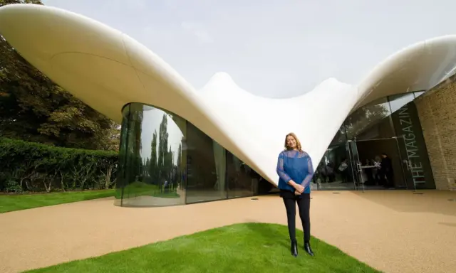Zaha Hadid steht vor einem ihrer ikonischen zaha hadid bauwerke, einem geschwungenen, weißen Dach mit Glasfront.