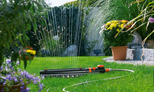Sistema de riego automático Gardena en césped verde, con flores amarillas y moradas al fondo.