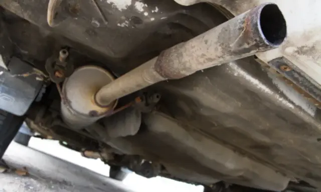 Vista inferior de un coche, mostrando el sistema de escape con un silenciador y tuberías oxidadas. El colector de escape es una parte crucial del sistema.