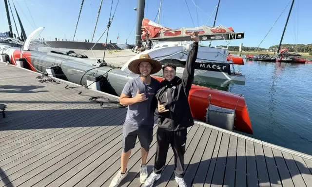 Deux jeunes hommes posent devant un bateau de course prêt pour une traversée atlantique. Le bateau est long, gris et rouge, avec des voiles partiellement visibles.