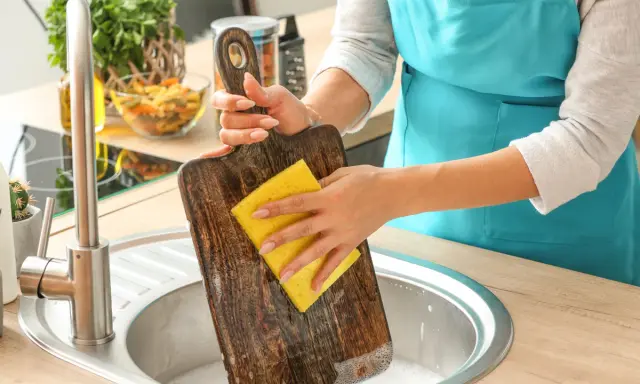 Frau reinigt ein Holz-Schneidebrett mit einem gelben Schwamm in der Spüle. Backpulver hilft, hartnäckige Flecken zu entfernen.