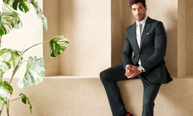 A man in a sharp green suit and black shirt poses confidently next to a lush plant.