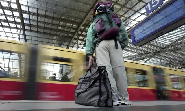 Traveler with backpack and duffel bag waits on a platform as a train speeds by.