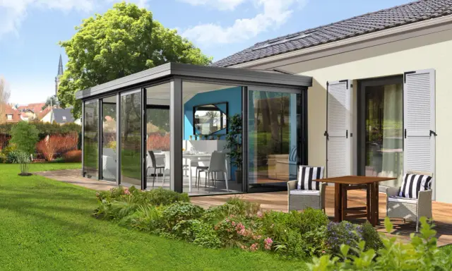 Moderne veranda sans permis de construire, avec table et chaises, offrant une vue sur un jardin verdoyant et des maisons.