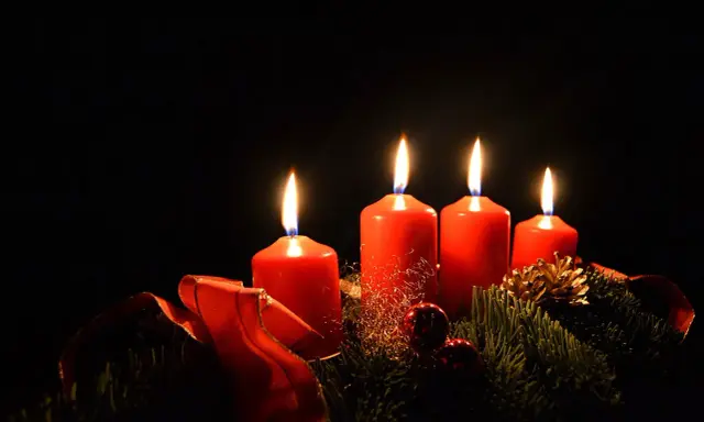 Fyra röda adventsljus brinner i mörkret, en del av en julkrans med granris och röda band. Detta är en visuell representation av vad betyder advent.