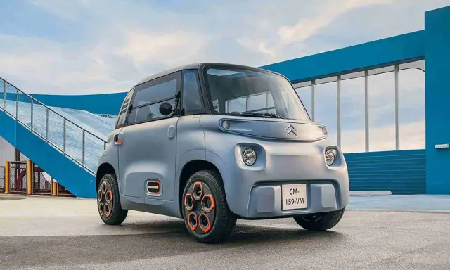 Un cuadriciclo pesado Citroën Ami gris con ruedas naranjas, aparcado frente a una estructura azul y una rampa.