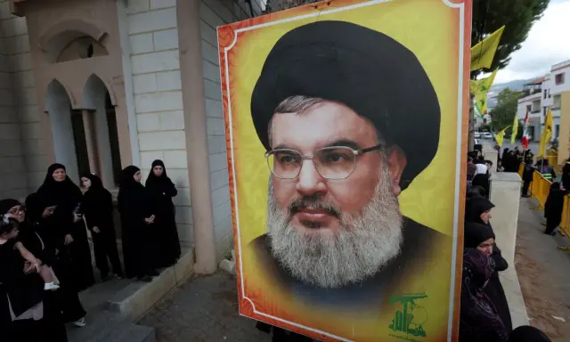 Wielki portret Hassana Nasrallaha, lidera Hezbollah, obok grupy kobiet w czarnych hidżabach.