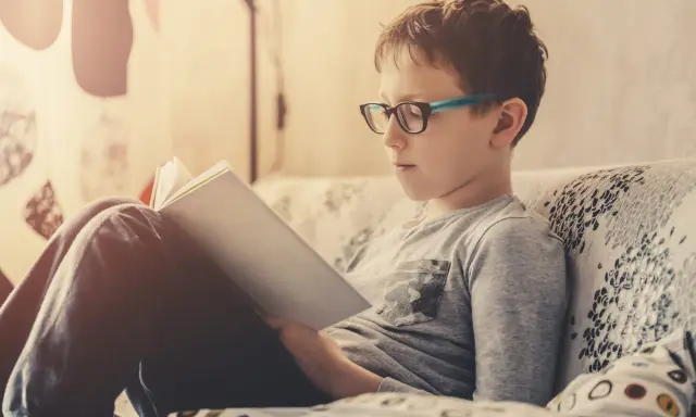 Bambino con occhiali legge un libro, concentrato su come imparare una poesia a memoria.