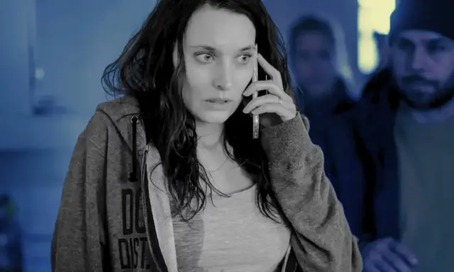 Przerażona kobieta rozmawia przez telefon, podczas gdy w tle widać zamazane postacie. Scena jak ze skandynawskich seriali kryminalnych.