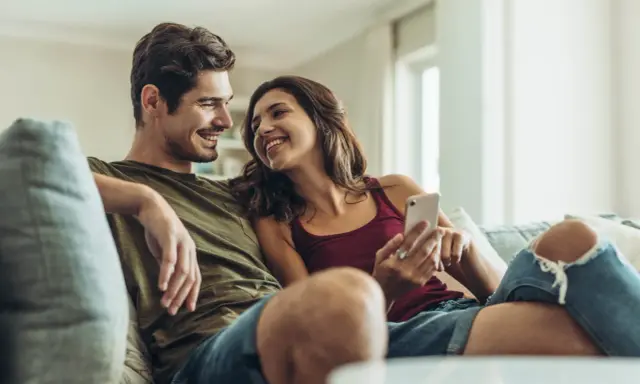 Pareja sonriente en el sofá, compartiendo un momento íntimo. Ella le muestra algo en su móvil, una forma de como expresar sentimientos.