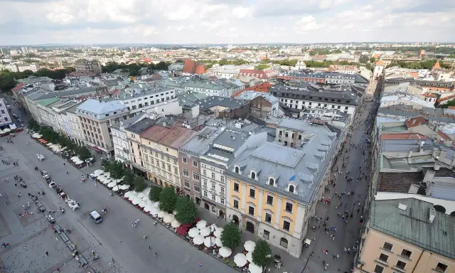 Widok z góry na Rynek Główny w Krakowie. Tłumy ludzi spacerują, a dorożki czekają na turystów. W tle widać zabytkowe kamienice i rozległą panoramę miasta.