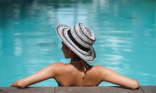 Donna con cappello a righe, braccia aperte, si gode il sole in piscina.