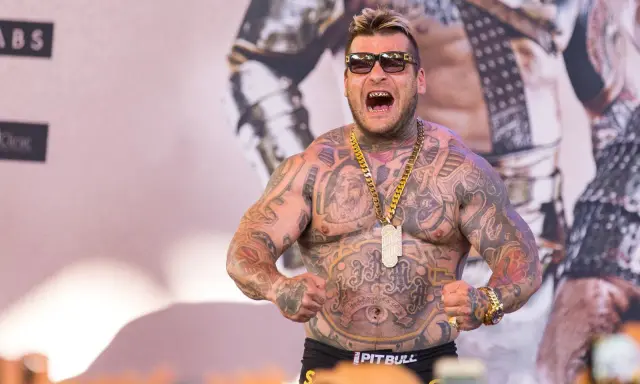 Popek kiedys, wytatuowany mężczyzna w okularach przeciwsłonecznych, z złotym łańcuchem i wisiorem, napina mięśnie.