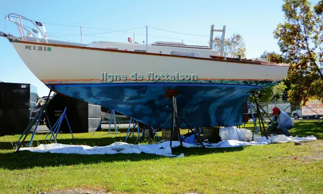 Un bateau est sur des chandelles, prêt pour une peinture. La ligne de flottaison est visible sur la coque bleue. Un homme travaille sur le bateau.