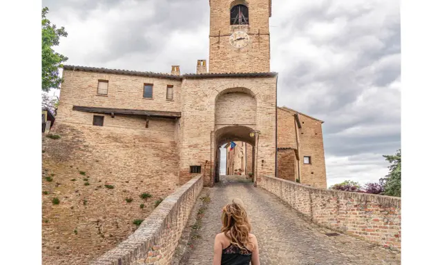 Passeggiata verso il borgo di Montegridolfo cosa vedere: un antico portale in mattoni con torre dell'orologio e un ponte acciottolato.
