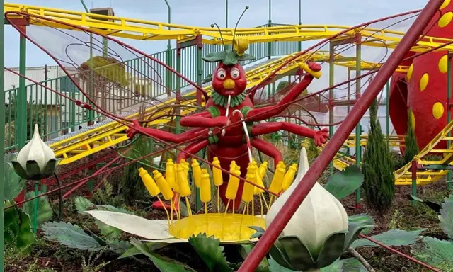 Eine rote Insektenfigur mit vielen Armen steht vor einer gelben Achterbahn.