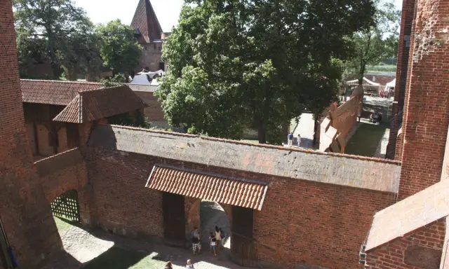Wielki zamek w Malborku, ceglane mury, dziedziniec z drzewami i ludźmi spacerującymi pośród zabytkowej architektury.