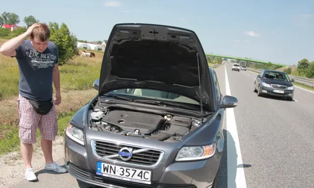 Mężczyzna zmartwiony przy otwartej masce Volvo, prawdopodobnie przegrzany silnik. W tle droga i pola.