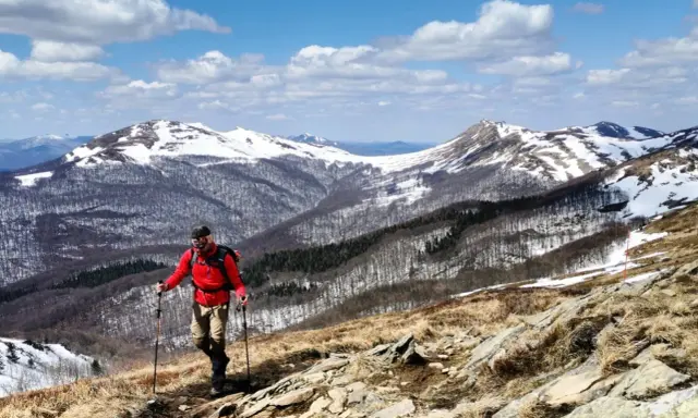 Mężczyzna w czerwonej kurtce uprawia nordic walking na górskim szlaku. W tle ośnieżone szczyty i las.