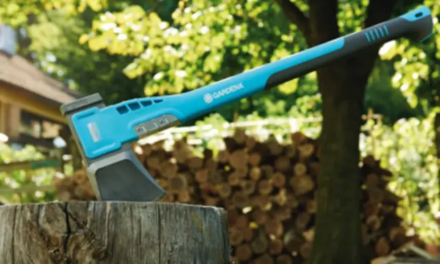 Une hachette bleue et grise est plantée dans un billot, prête à couper du bois. Une pile de bois est visible en arrière-plan.