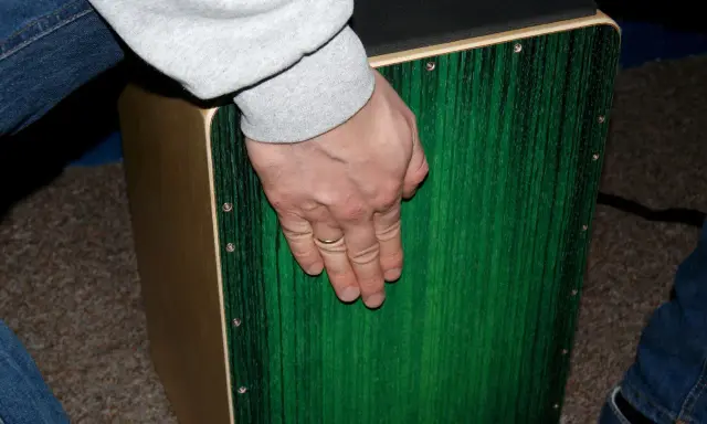 Dłoń z obrączką na palcu spoczywa na zielonym pudle perkusyjnym.