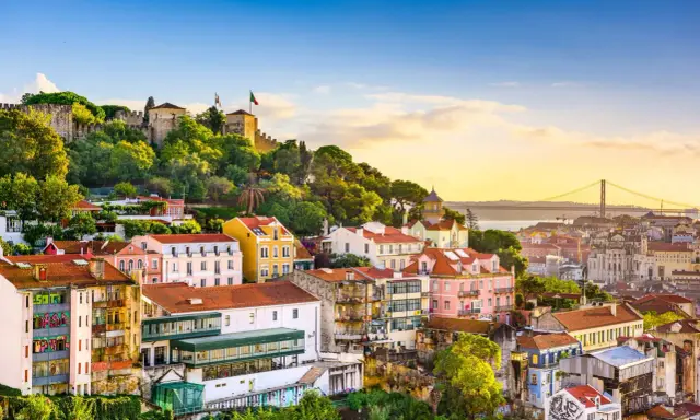 Bunte Häuser und eine Burg auf einem Hügel in Lissabon, der Hauptstadt Portugals. Im Hintergrund die Hängebrücke über den Tejo.