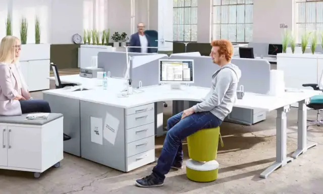 Un espace de travail open space moderne avec des bureaux modulables, des plantes vertes et des sièges colorés. Deux personnes discutent, une troisième travaille sur son ordinateur.